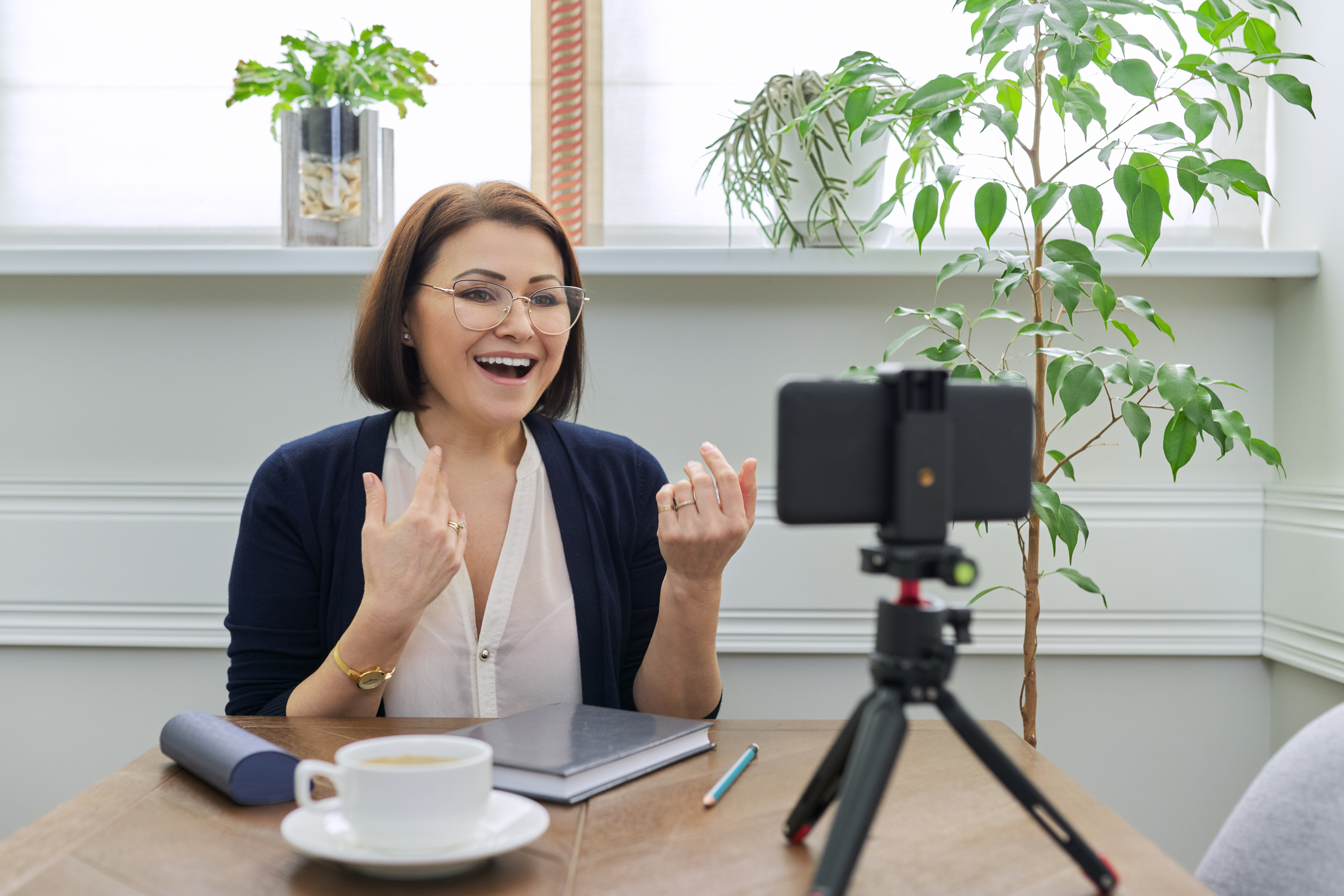 Video bodytalk online, woman looking at smartphone webcam on tripod, recording video, talking online, sitting at table at home.